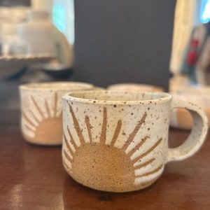 May include: A white ceramic mug with a brown speckled glaze and a white sun design. The mug has a handle and is perfect for coffee or tea.