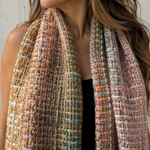 May include: A woven scarf featuring a textured grid pattern in a multi-colored palette of soft pink, muted orange, earthy brown, and bright turquoise. The fabric displays a dense, interlaced construction with visible yarn loops throughout the design.