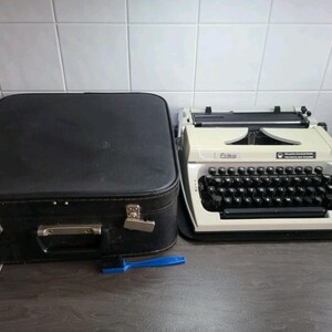 May include: An off-white Erika typewriter with black keys and a black carrying case. A blue cleaning brush is next to the typewriter. The typewriter has the brand name "Erika" on it.