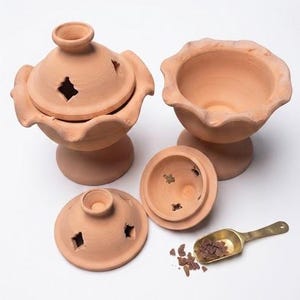May include: Two terracotta incense burners with scalloped edges and matching lids featuring star-shaped cutouts. One burner is assembled, while the other is shown with its lid removed. A small brass scoop contains dark resin incense pieces.
