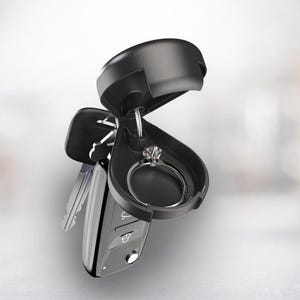 May include: A black car key fob with a hidden compartment holding a diamond ring. The key fob is open, revealing the ring. The ring has a silver band and a large, clear gemstone. Other keys are attached.