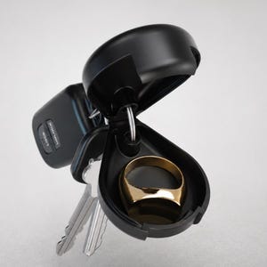 Könnte beinhalten: Ein schwarzer Schlüsselanhänger mit einem offenen Fach, das einen goldenen Ring enthält. Weitere Schlüssel sind am Schlüsselanhänger befestigt. Der Hintergrund ist schlicht weiß.