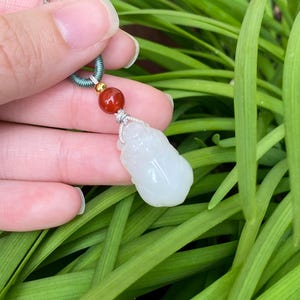 May include: A white jade pendant in the shape of a small Buddha figure, suspended from a gray cord with a teal and gold accent. A red bead is also included in the design. The pendant is held against a backdrop of green foliage.