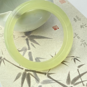 May include: A pale green jade bangle bracelet. The smooth, circular bracelet is set against a backdrop of a white ceramic bowl and a paper with a bamboo design. The jade has a translucent quality.