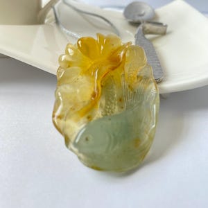May include: A translucent pendant featuring a carved fish design in shades of pale green and amber yellow. The pendant is suspended from a thin silver-toned chain and displays intricate detailing on the fish scales and fins within the stone.