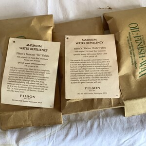 以下が含まれることがあります： 茶色の紙袋がいくつかあり、製品情報タグが付いています。タグには「Maximum Water Repellency」と「Oil Finish Wax」と書かれています。テキストは生地の耐水性と耐久性について説明しています。「Filson」のブランド名が見えます。