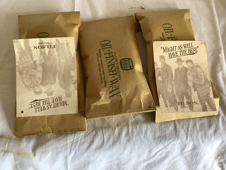 以下が含まれることがあります： Filsonオイルフィニッシュワックスの茶色の紙パッケージがいくつかあります。各パッケージには、「Might as well have the best」というテキストとFilsonのロゴが入ったヴィンテージスタイルのラベルが付いています。パッケージは白い表面に配置されています。