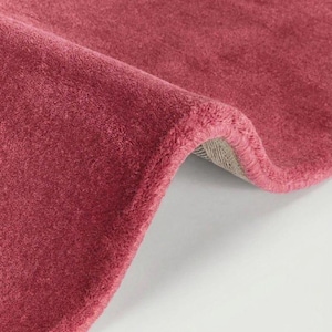 May include: A solid deep pink rug with a soft, plush texture. The rug is folded over to reveal a light-colored, woven backing material along the curved edge. The surface of the rug features a dense, uniform pile that creates a consistent color.