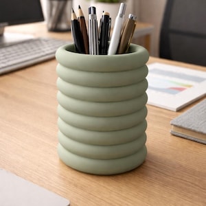 Op de afbeelding: Een saliegroene pennenhouder met een uniek ontwerp van gestapelde ringen. De houder bevat verschillende pennen en potloden, waaronder zwarte, bruine en zilveren pennen. De pennenhouder staat op een houten bureau, met een computertoetsenbord en andere kantoorbenodigdheden op de achtergrond.