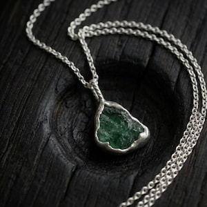 Puede incluir: Un collar de plata con un colgante con una piedra preciosa verde esmeralda en bruto. El colgante está encerrado en un engaste de plata y cuelga de una delicada cadena de plata. El collar se exhibe sobre una superficie oscura y texturizada.