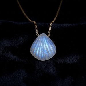 May include: A gold chain necklace with a shell-shaped pendant. The pendant has a pearlescent, blue and white glow, with detailed ridged lines. The necklace is set against a dark background.