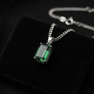 May include: A silver necklace with a rectangular, emerald-green gemstone pendant. The pendant is set in a silver frame and hangs from a silver chain. The necklace is displayed on a black surface.