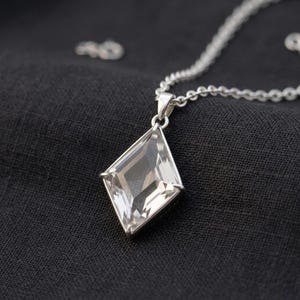 Peut inclure: Un collier en argent avec un pendentif clair en forme de losange. Le pendentif est facetté, captant la lumière. La chaîne est fine, et le pendentif est la pièce maîtresse sur un fond sombre. Un bijou.