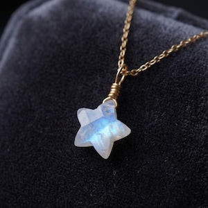 May include: A delicate gold chain necklace featuring a star-shaped pendant. The pendant is a pale blue and white gemstone, with a faceted surface. The necklace is displayed against a dark background.