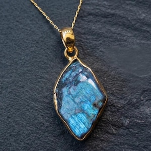 May include: A gold-colored necklace featuring a pendant with a blue and teal gemstone. The gemstone is irregularly shaped and set in a gold bezel. The necklace is displayed on a dark gray surface.