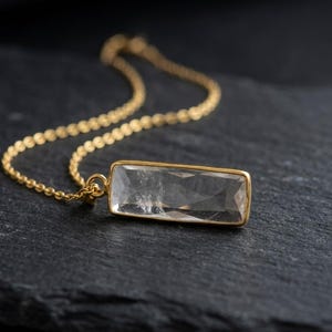 May include: A gold chain necklace featuring a rectangular, faceted clear crystal pendant set in a gold frame. The pendant is elongated and horizontally oriented, showcasing a transparent, geometric cut that reflects light across its surface.
