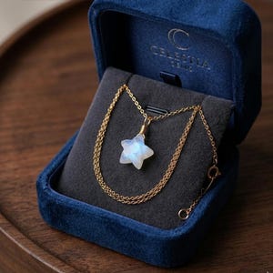 May include: A gold chain necklace with a star-shaped pendant, presented in a blue velvet jewelry box. The pendant is a light blue and white gemstone. The jewelry box has the text "CELESTIA GEMS" printed on the inside of the lid.