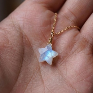 May include: A delicate gold necklace featuring a star-shaped pendant. The pendant is crafted from a translucent, iridescent stone, displaying shades of blue and white. The necklace chain is a fine, gold-colored link.
