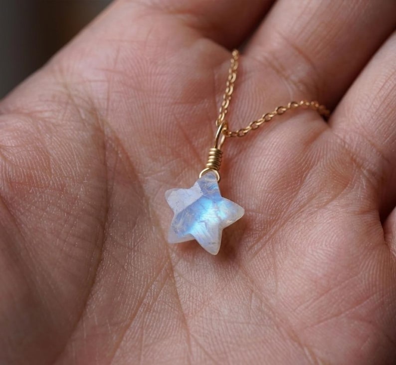 May include: A delicate gold necklace featuring a star-shaped pendant. The pendant is crafted from a translucent, iridescent stone, displaying shades of blue and white. The necklace chain is a fine, gold-colored link.