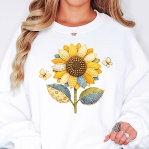 May include: White sweatshirt featuring a large sunflower design with yellow, blue, and green patchwork details. Two small butterflies are on either side of the flower. The sunflower has a brown center and a stem with leaves.