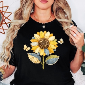 May include: Black t-shirt featuring a large sunflower design with yellow, blue, and green patchwork details. Two small butterflies are on either side of the flower. The shirt has a round neckline.