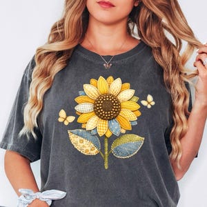 May include: Dark gray t-shirt featuring a large sunflower design with yellow, blue, and green patchwork details. Two yellow butterflies flank the flower. The shirt has a relaxed fit and short sleeves.