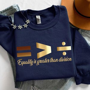 May include: Navy blue sweatshirt with gold text that reads "Equality is greater than division." The design includes mathematical symbols. A silver necklace with flower-shaped pendants and a brown hat are also visible.