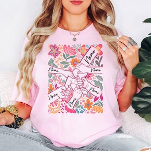 May include: Pale pink t-shirt with a floral design and hands holding each other. Each hand has the word "Name" written on it. The shirt has a crew neck and short sleeves. The floral design includes pink, orange, and blue flowers.