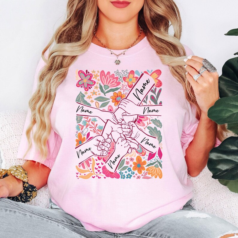 May include: Pale pink t-shirt with a floral design and hands holding each other. Each hand has the word "Name" written on it. The shirt has a crew neck and short sleeves. The floral design includes pink, orange, and blue flowers.