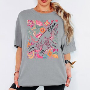 May include: Grey t-shirt with a floral design and hands holding each other. The names "Emma", "Steve", "Olivia", and "Liam" are written on the design. The floral design includes pink, orange, and blue flowers.