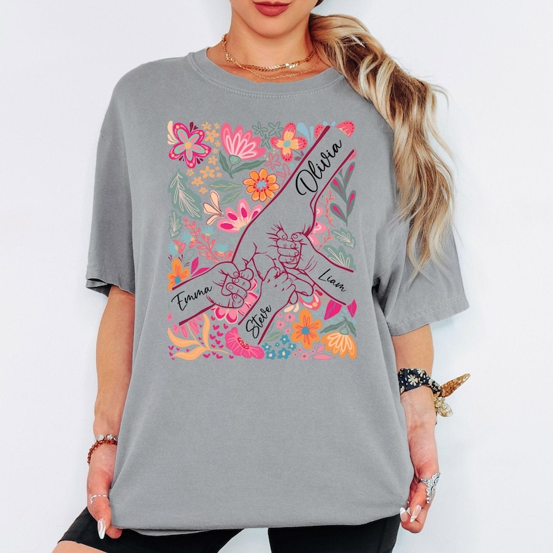 May include: Grey t-shirt with a floral design and hands holding each other. The names "Emma", "Steve", "Olivia", and "Liam" are written on the design. The floral design includes pink, orange, and blue flowers.