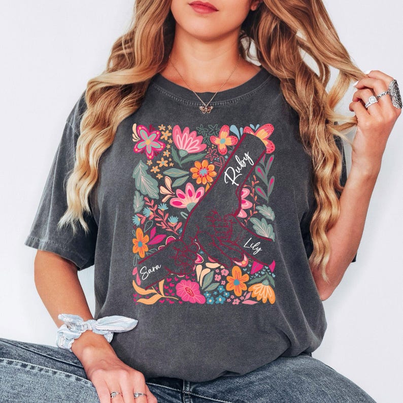 May include: Dark grey t-shirt with a floral design and the names "Ruby", "Lily", and "Sara". The design features colourful flowers in pink, orange, and blue. The shirt has short sleeves and a crew neck.