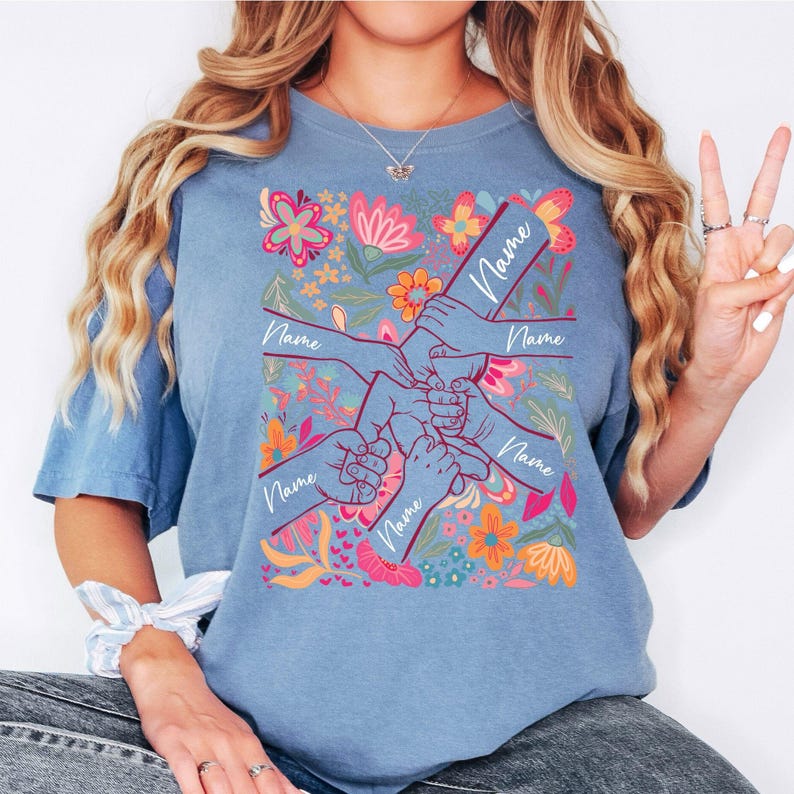 May include: Pale blue t-shirt featuring a floral pattern and a graphic of hands clasped together. Each arm in the graphic has the word "Name". The shirt has a crew neck and short sleeves.