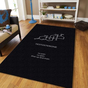 May include: Black rectangular rug featuring a white graphic of the testosterone molecule and the word "TESTOSTERONE". Below the molecule are the words "no pain no gain Shat up and train". The rug is placed on a hardwood floor.