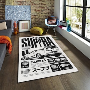 May include: A rectangular white rug with a black graphic of a Toyota Supra car. The rug features the word "SUPRA" in large black letters, along with technical details and Japanese text. The rug is placed on a wooden floor.