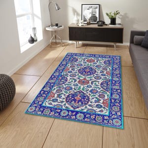 Op de afbeelding: Rechthoekig vloerkleed met een gedetailleerd bloemenpatroon in blauw, rood en groen op een witte achtergrond. Het kleed heeft een blauwe rand en ligt op een lichte houten vloer. Het ontwerp bevat ingewikkelde bloemen- en geometrische elementen.