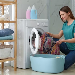 May include: A woman loads a washing machine with colorful clothing. A light blue laundry basket sits in front of the machine. A wooden shelf holds towels and baskets. Bottles of laundry detergent are on top of the washing machine.