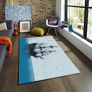 May include: A rectangular area rug featuring a nautical design. The rug has a light blue background with a dark blue and black illustration of a tall ship. A vibrant blue section represents the sea. The rug is placed on a wooden floor.