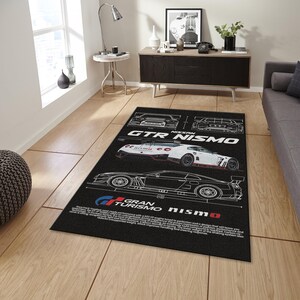 May include: Black rectangular area rug featuring a white outline design of a Nissan GTR Nismo race car. The rug includes the text "GTR Nismo" and "Gran Turismo Nismo" in white and red. The rug is placed on a wooden floor.