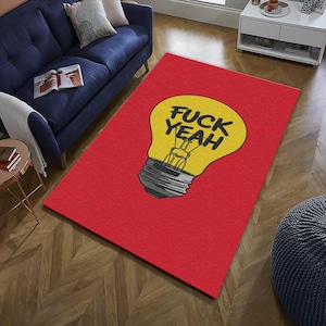 Könnte beinhalten: Ein roter rechteckiger Teppich mit einer großen gelben Glühbirnen-Grafik. Der Text "FUCK YEAH" ist in Schwarz auf der Glühbirne zu sehen. Der Teppich liegt auf einem Holzboden, mit einem blauen Sofa und einem Beistelltisch im Hintergrund.