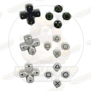 May include: A collection of replacement controller buttons in black and silver. The image shows directional pads and individual buttons with symbols like triangles, squares, circles, and X's. The buttons are for gaming consoles.