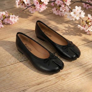 May include: A pair of black leather split-toe ballet flats with small bows. The flats have a unique design with a separate section for the big toe. The word "Runabi" is printed inside the shoe. Cherry blossoms are in the background.