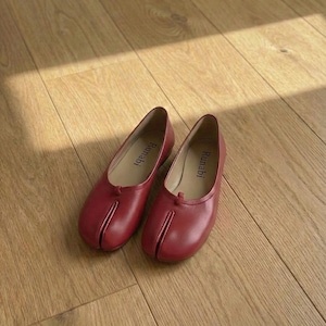 Op de afbeelding: Een paar rode leren ballerina's met een split teenontwerp. De schoenen worden gepresenteerd op een lichtbruine houten vloer. De merknaam "Runabi" staat op de binnenzool van elke schoen.