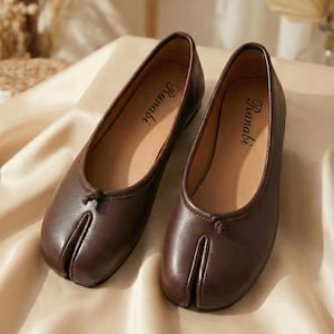 Puede incluir: Un par de bailarinas de cuero marrón con diseño de puntera dividida. Los zapatos tienen un pequeño lazo en la parte superior y la palabra "Runabi" impresa en la plantilla. Se muestran sobre una superficie beige.