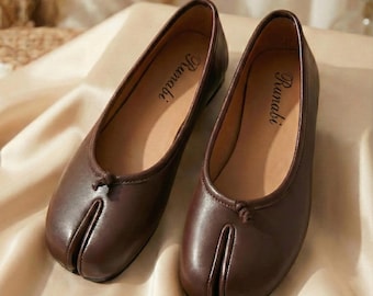 May include: A pair of brown leather ballet flats with a split-toe design. The shoes have a small bow detail at the top and the word "Runabi" printed on the insole. The flats are displayed on a beige surface.