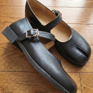 May include: A pair of black leather Mary Jane shoes with a Tabi toe design. Each shoe features a strap with a silver buckle. The shoes are on a wooden surface. The word "runabi" is printed on the insole.