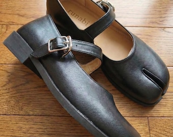 May include: A pair of black leather Mary Jane style shoes with a Tabi toe design. Each shoe has a strap with a silver buckle. The shoes are on a wooden surface. The word "runabi" is printed on the insole.