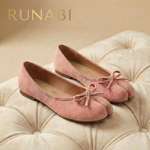 Op de afbeelding: Een paar roze ballerina's met een split-teenontwerp en kleine strikjes. De schoenen zijn gemaakt van een zacht, suède-achtig materiaal en hebben de merknaam "Runabi" aan de zijkant. Ze worden gepresenteerd op een gecapitonneerd, crèmekleurig oppervlak.