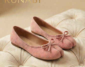 May include: A pair of pink ballet flats with a split-toe design and small bows. The shoes are made of a soft, suede-like material and have the brand name "Runabi" printed on the side. They are displayed on a tufted, cream-colored surface.
