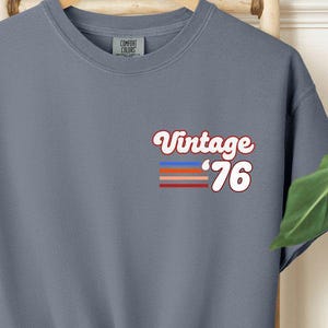 May include: A dusty blue t-shirt with the text "Vintage '76" in a retro font. The graphic includes horizontal stripes in red, blue, and orange. The shirt is knotted at the bottom.